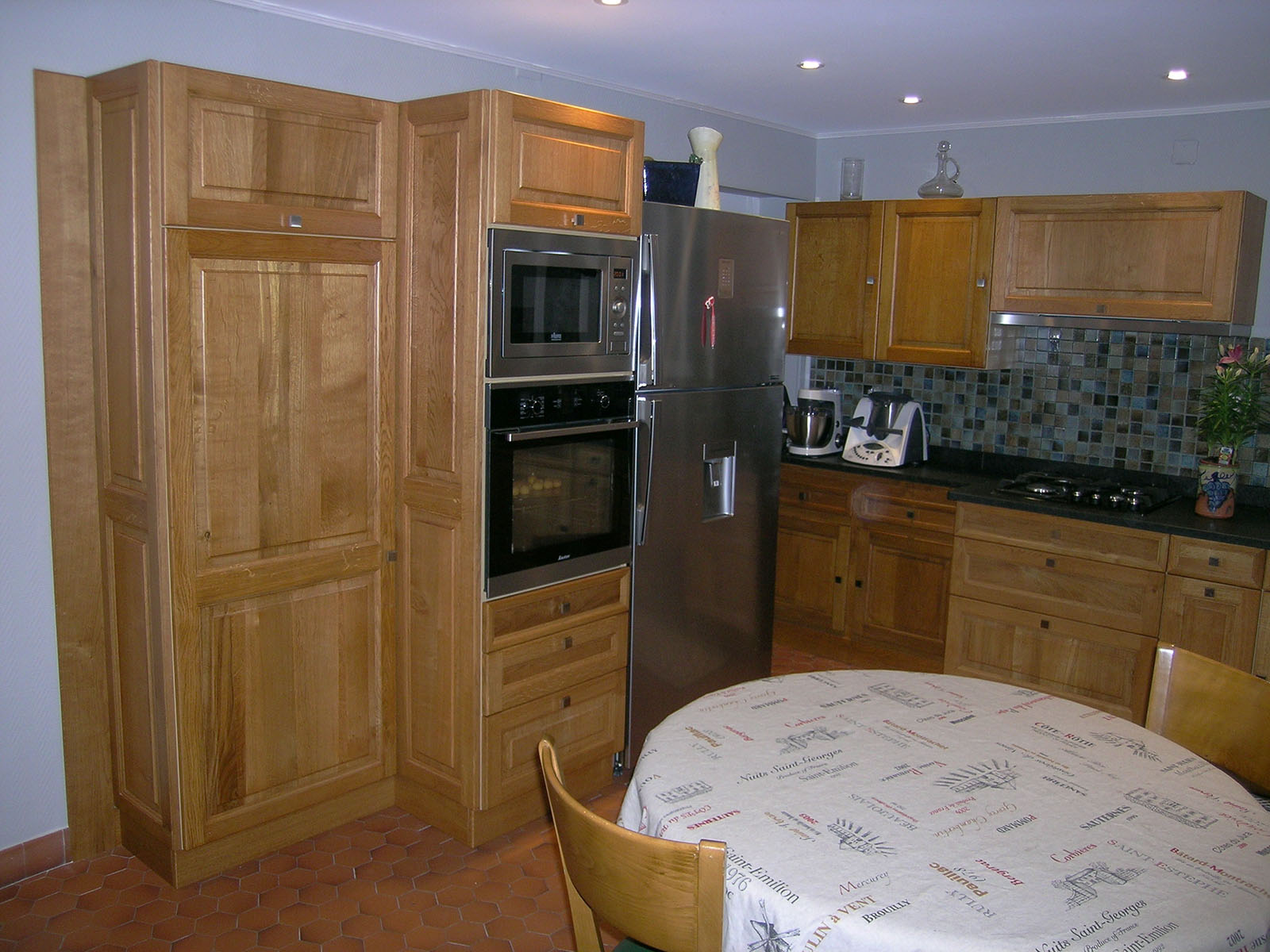 Façade — Chêne massif, teinté, verni.
Poignée — Bouton carré inox.
Plan de travail — Granite noir.
Electroménager — Micro-onde, four, frigo, plaque de cuisson, hotte encastrée.